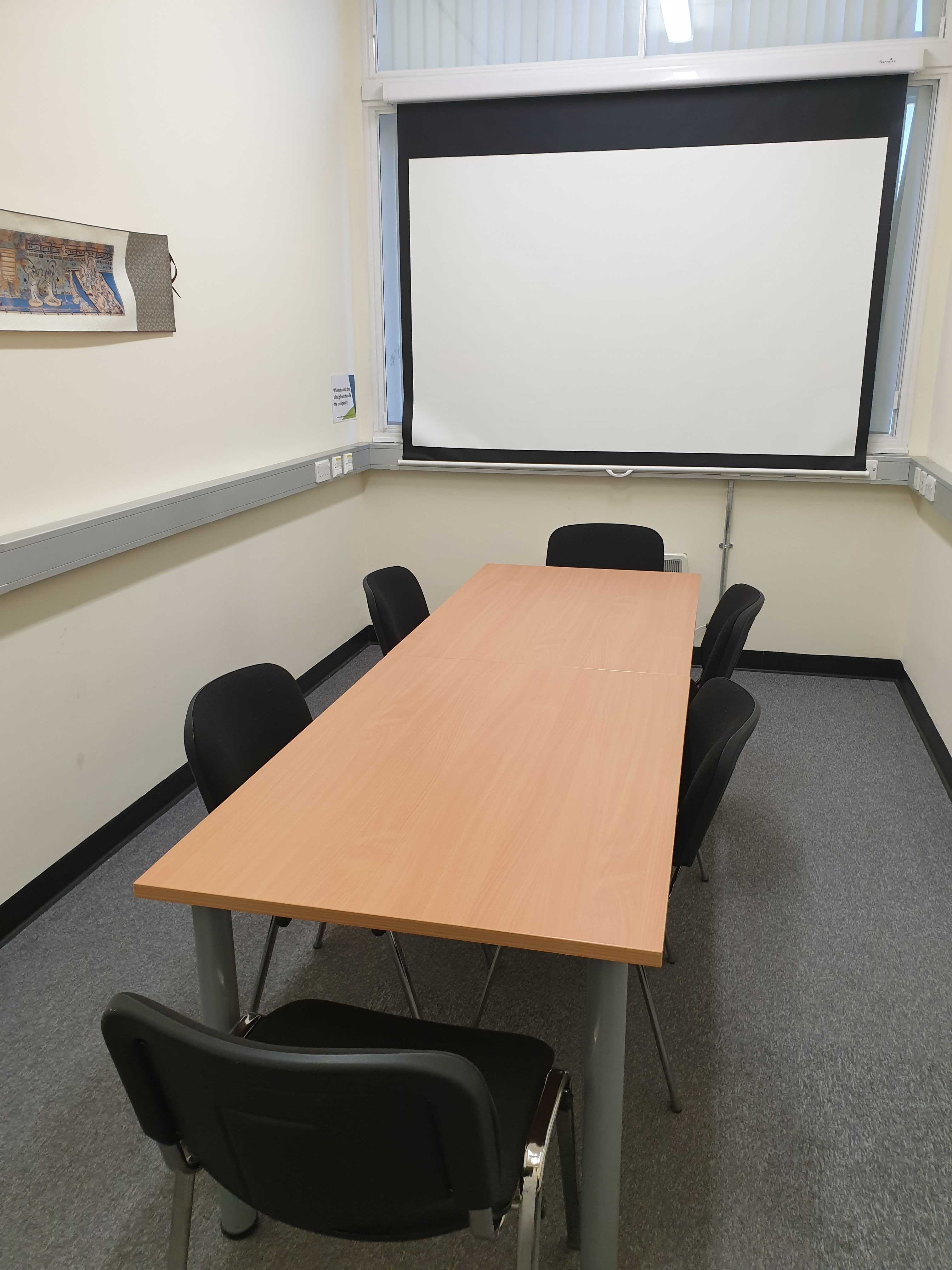 Photograph showing the standard layout of the Biomedical Campus Committee Room - a room with six chairs around a large boardroom table, with a projector screen on the end wall.