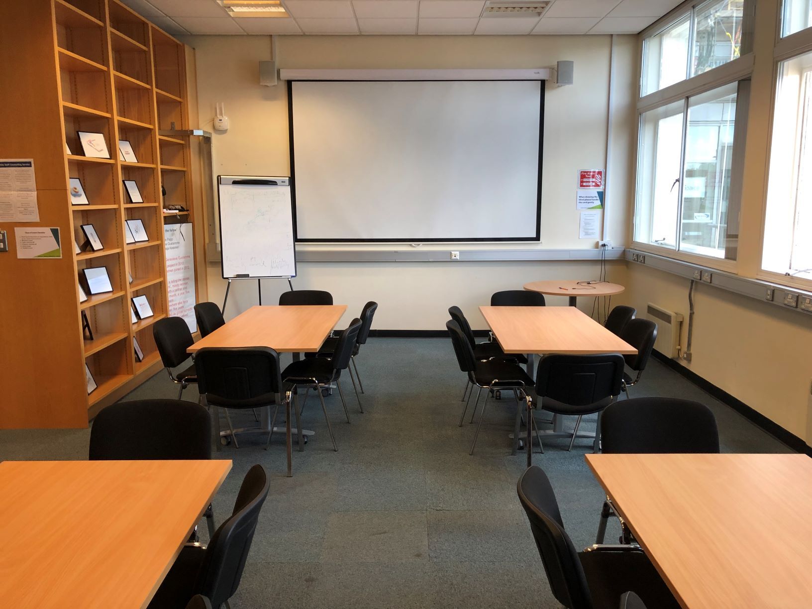 Photograph showing the standard layout of the Newman Library - a room with four rectangular tables, each with six chairs, with a projector screen on the end wall.