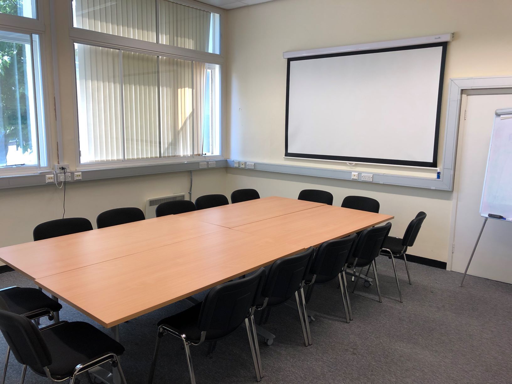 Photograph showing the standard layout of the Biomedical Campus Seminar Room - a room with twelve chairs around a large boardroom table, with a projector screen on the end wall.