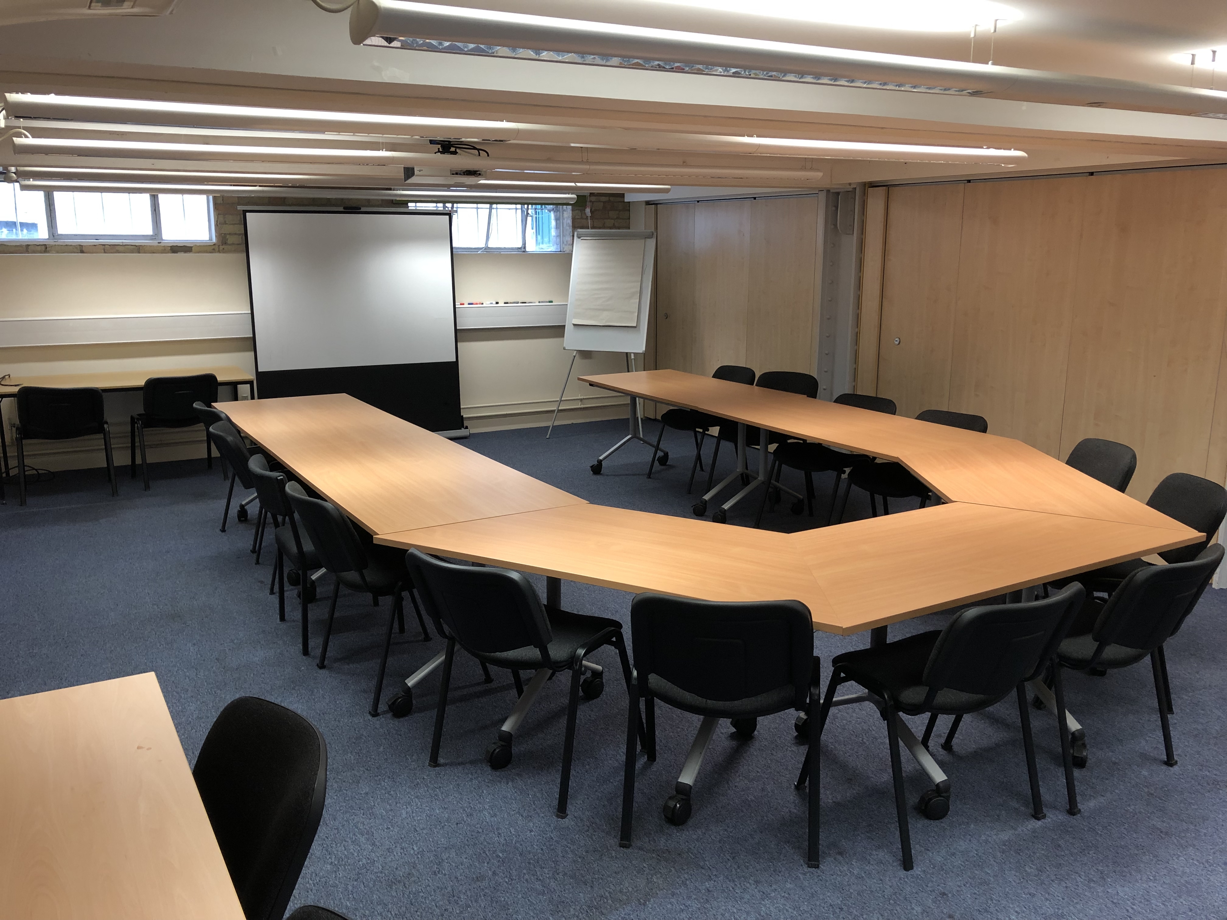Photograph showing the standard layout of the Mill Lane Seminar Room - a room with fourteen chairs around a large boardroom table, with a projector screen on the end wall.
