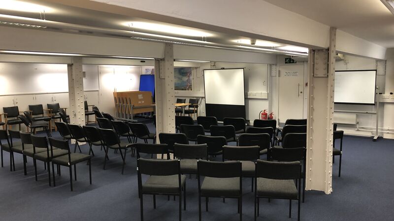 Photograph showing the lecture-style layout of the Eastwood Room - a room with five rows of chairs facing a projector screen on the end wall.