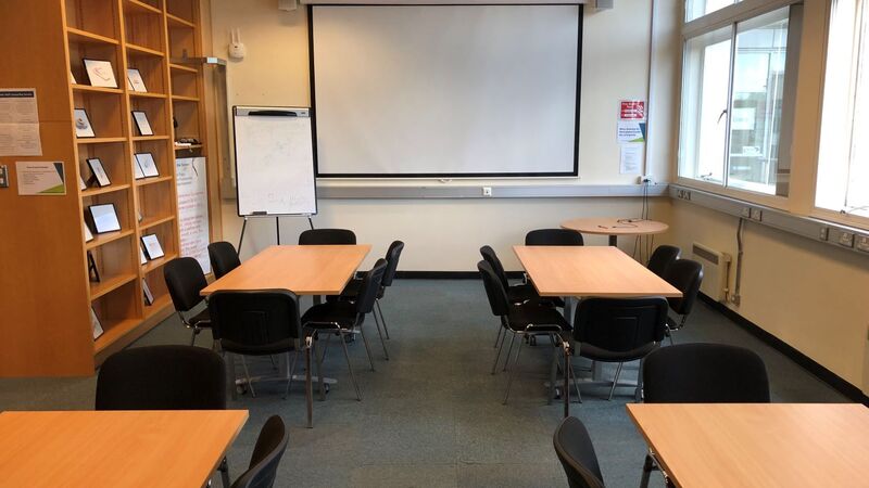 Photograph showing the standard layout of the Newman Library - a room with four rectangular tables, each with six chairs, with a projector screen on the end wall.