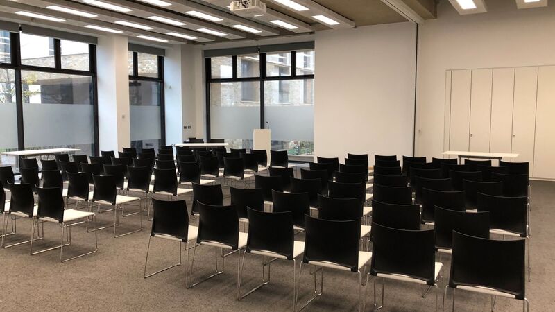 Photograph showing the lecture-style layout of Sanders Hall - a room with seven rows of chairs facing a lectern and projector screen on the end wall.