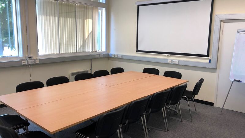Photograph showing the standard layout of the Biomedical Campus Seminar Room - a room with twelve chairs around a large boardroom table, with a projector screen on the end wall.