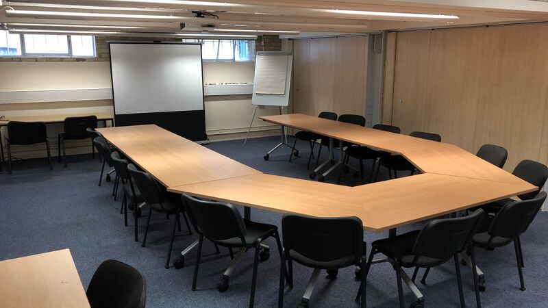 Photograph showing the standard layout of the Mill Lane Seminar Room - a room with fourteen chairs around a large boardroom table, with a projector screen on the end wall.