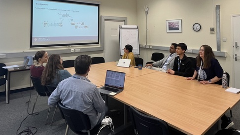 The Journal Club hold a meeting, gathered around a table while looking at a large screen at the front of the room
