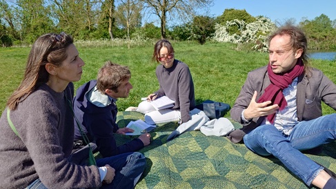 Four people sit on a picnic blanket beside the river Cam on a sunny day. They are deep in conversation.
