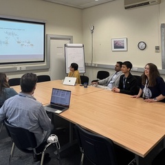 The Journal Club hold a meeting, gathered around a table while looking at a large screen at the front of the room