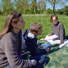 Four people sit on a picnic blanket beside the river Cam on a sunny day. They are deep in conversation.