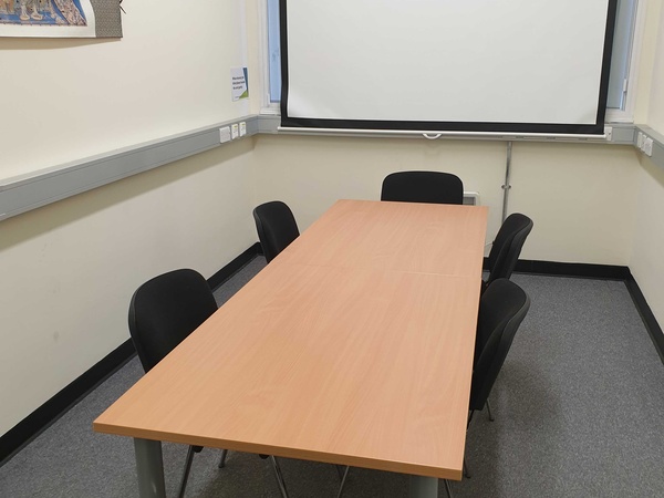 Photograph showing the standard layout of the Biomedical Campus Committee Room - a room with six chairs around a large boardroom table, with a projector screen on the end wall.